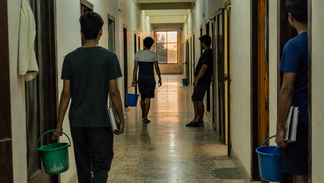 A hostel corridor seen from one end with students from behind carrying buckets and books, doors open, evening light through the far window.