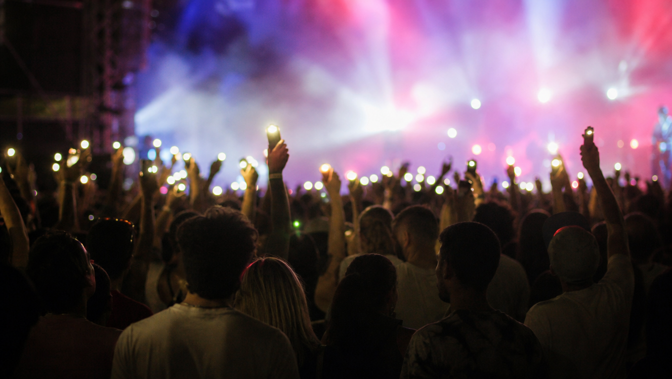 A concert crowd seen from behind holding up phone lights toward a stage washed in colour, hands raised, haze.