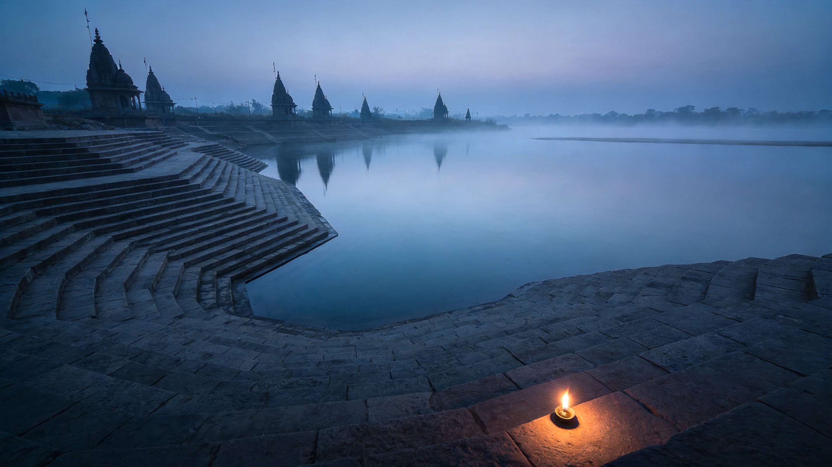 Tiered stone river ghats at blue hour with mist on the water and shrine spires on the far bank.