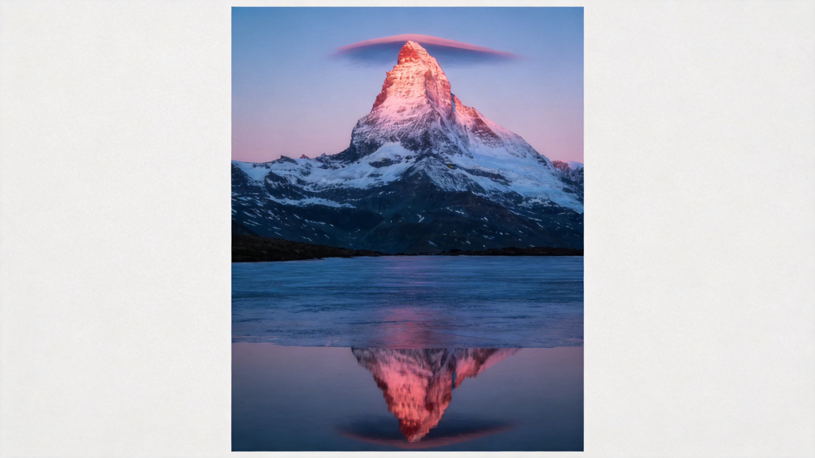 Solitary symmetrical snow peak lit pink by first light above a dark valley and a mirroring frozen lake.