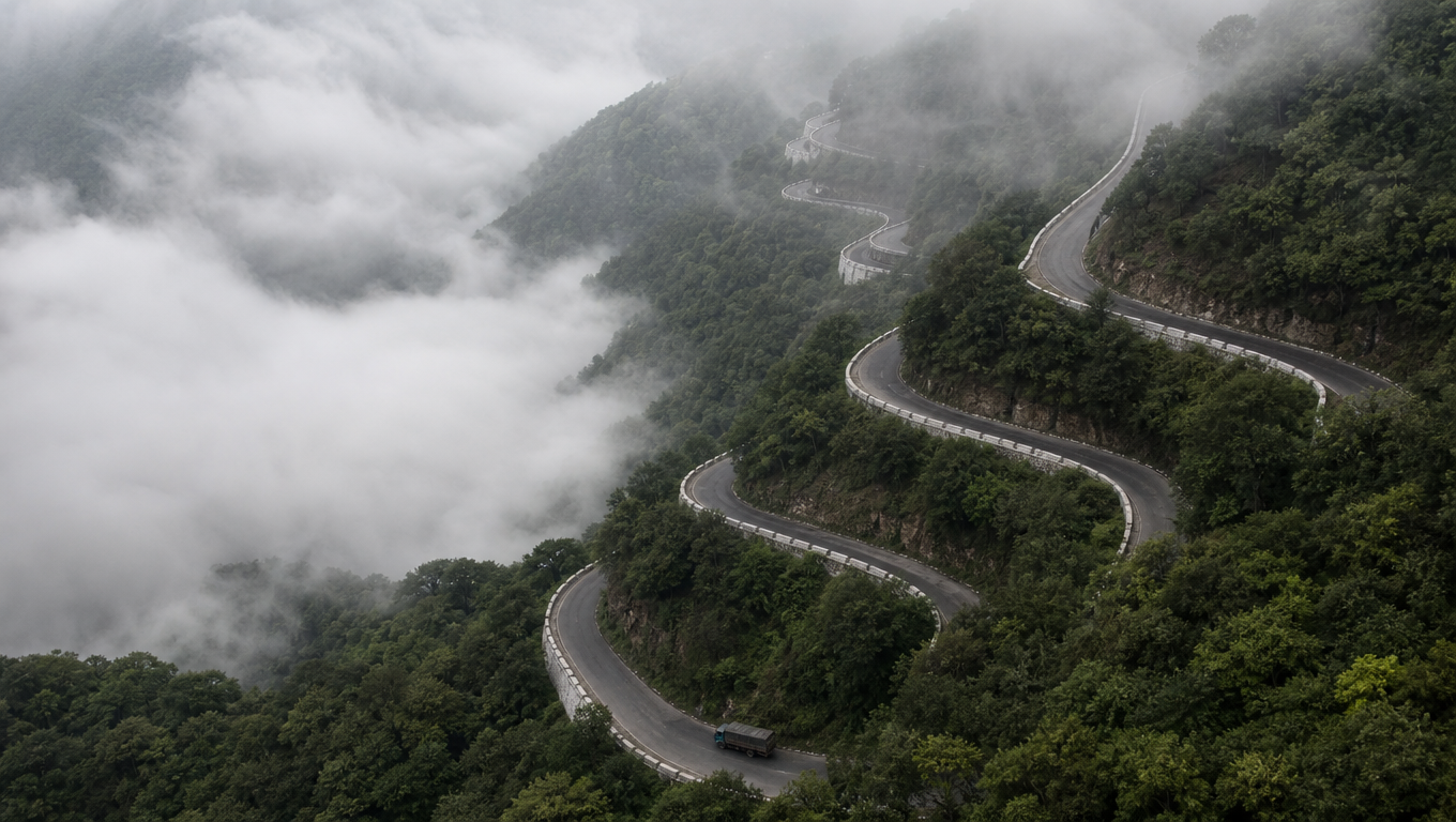 Stacked hairpin bends of a mountain ghat road climbing a forested slope through valley mist.