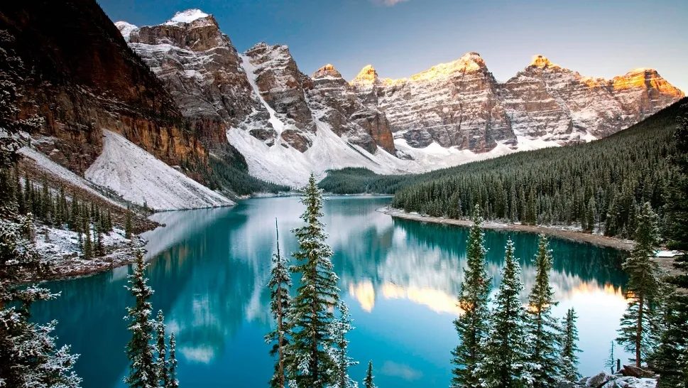 Turquoise glacial lake mirroring snow-dusted mountain peaks at sunrise, framed by dark spruce forest.