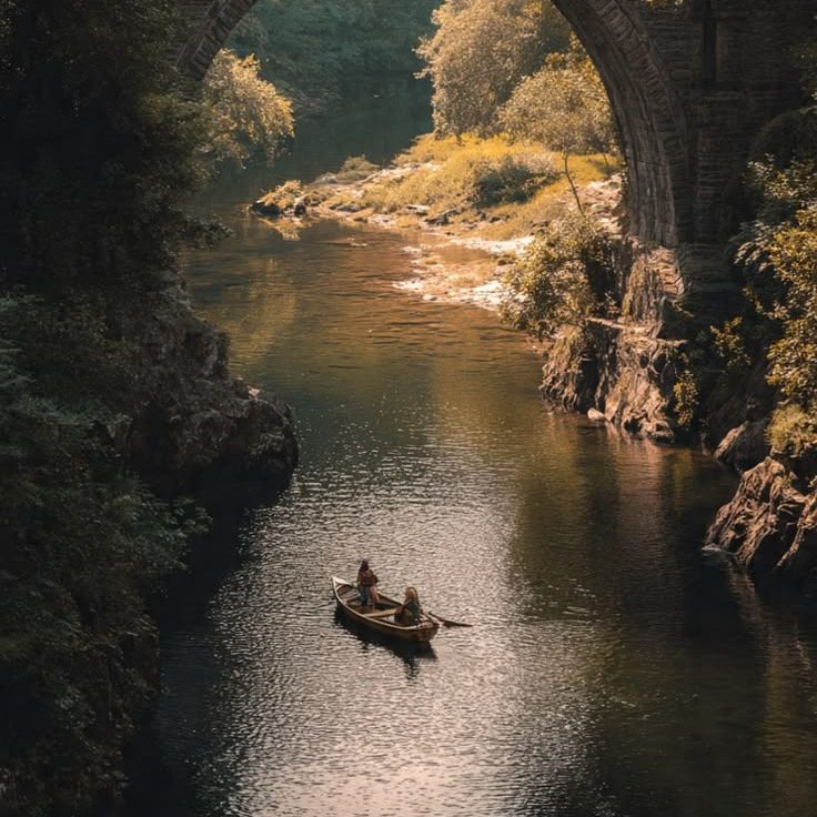 Two people rowing a small boat along a wooded river beneath the arch of an old stone bridge.